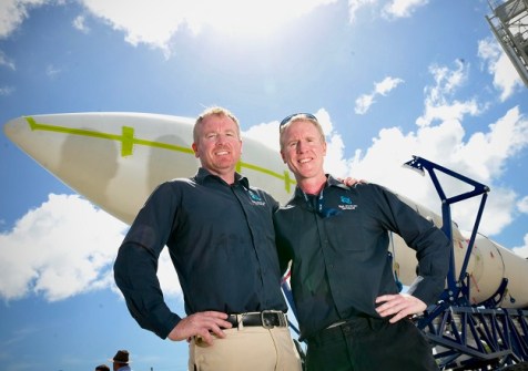 Two men standing side by side, with a rocket laid out horizontally behind them.