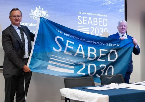 Two men holding between them a flag, upon which are the words Seabed 2030, during the ocean floor mapping agreement signing.