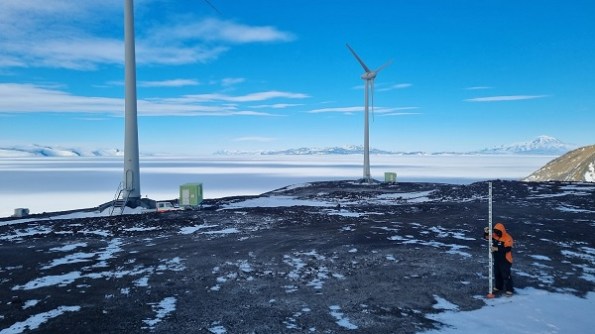 A rocky area in Antarctica, with two large wind turbine structures and a surveyor making measurements in the foreground.