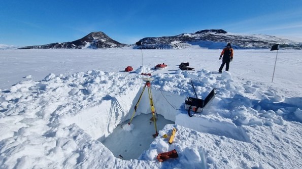 A rectangular hole dug in the ice in Antarctica, with a person standing nearby and surveying equipment arranged in places.