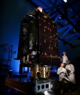 A GPS satellite inside a clean room, with three people standing next to it.