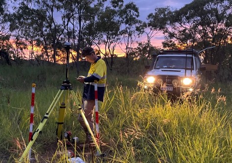 A man standing in grassland at dusk, conducting mapping and surveying, illuminated by the headlights of a 4WD vehicle behind him