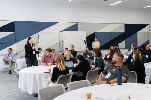 A scene from a recent Locate conference, showing people seated at round tables listening to a person standing, holding a microphone and speaking
