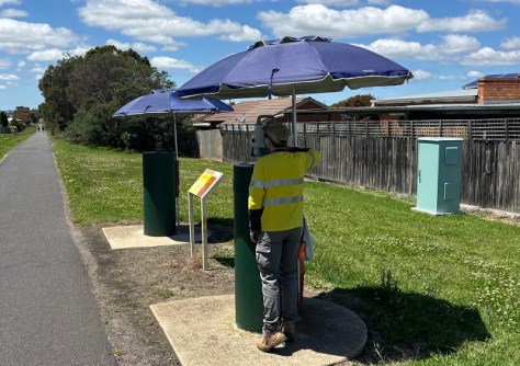 A surveyor standing next to a baselines calibration pillar, looking through a surveying device and using an umbrella to shade himself from the sunlight