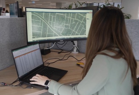 A woman sitting at a desk and looking at a computer screen, on which the Land iQ platform is displaying an overhead map of a suburb