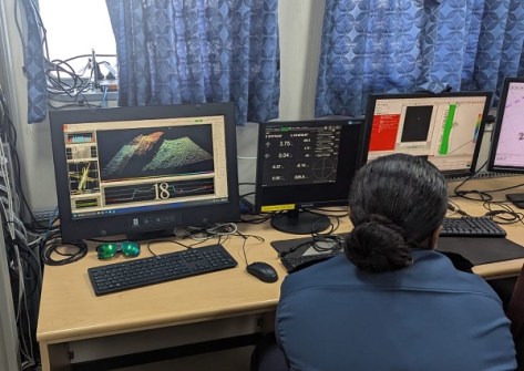 A view over the shoulder of a woman sitting at a desk looking at computer screens, aboard the RFNS Volasiga