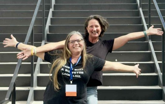 Two smiling women, one standing behind the other, with their arms outstretched, at the Women Surveyors Summit