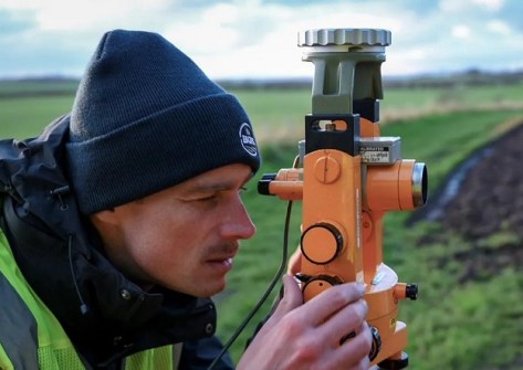 A close up of a man looking through a surveying instrument mounted on a tripod while investigating the three norths