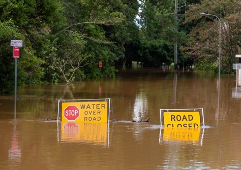A road flooded brown water, with half-submerged signs saying 'Road closed' and 'Water over road', with trees in the background.