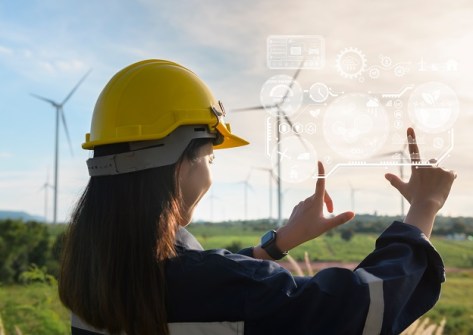 A woman in a yellow hardhat, holding her forefingers up in front of her as if measuring something, with wind turbines and sunset in the background. To represent women in GIS.