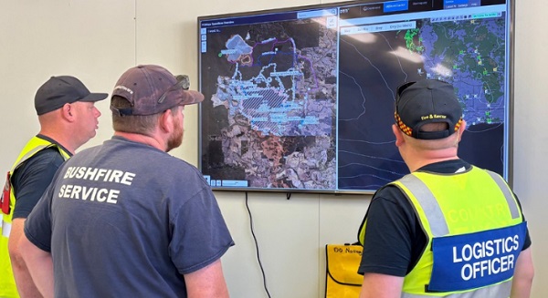 Three men with their backs to the camera, looking at a wall-mounted computer screen on which are depicted bushfire management maps