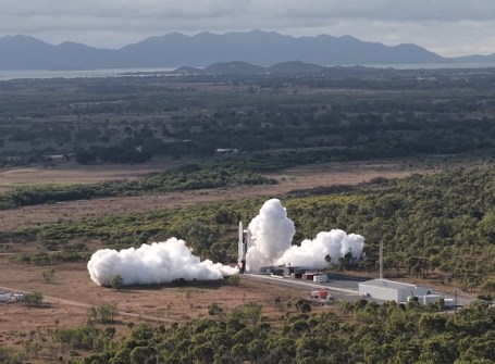 A distant view of a Gilmour Space rocket launch, which white plumes of smoke coming out to the left and the right at the base of the rocket.