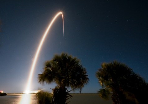 A nighttime GPS III satellite launch, showing a glowing arc of light from the launch vehicle motors rising from the launch pad and ascending into space.