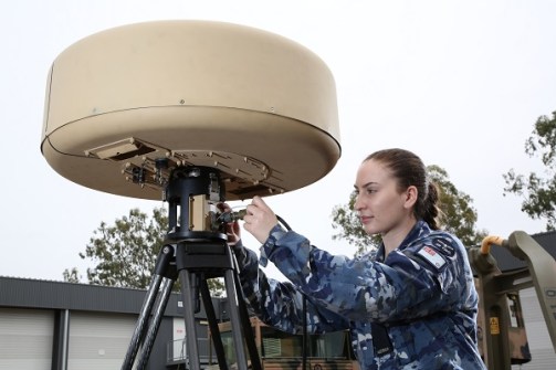 A woman in RAAF uniform connecting a cable to a satellite antenna, which is a squat cylinder about 50cm tall, mounted on a sturdy tripod.
