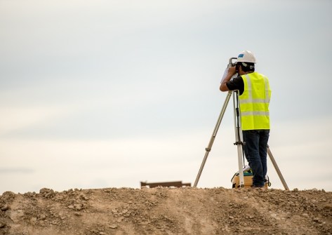 A surveyor in the construction industry, standing on a mound of dirt and looking through a surveying instrument