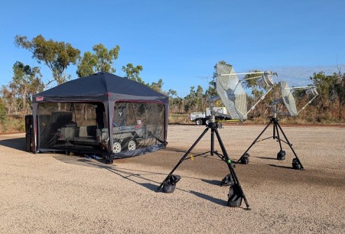 In a cleared bush setting, two small satellite dishes are on the right and a trailer with equipment is situated on the left within a tent.