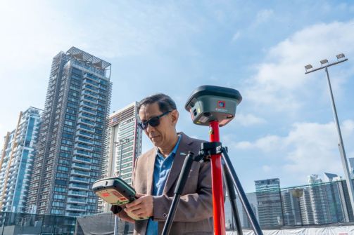 Dr Victor Khoo, the new Chief Surveyor of Singapore, using surveying equipment outside with tall buildings and blue sky in the background