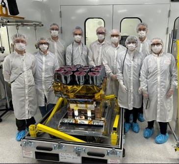 Nine people wearing gowns and masks, stand next to an EarthDaily satellite in a cleanroom