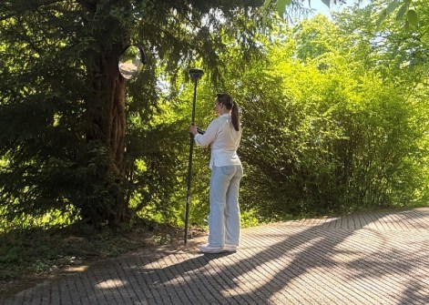 Phoebe Davis, standing on a path amongst trees, holding a GNSS rover on a pole