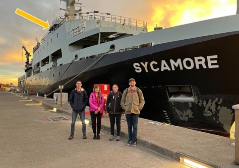 Four people standing on a dock with a large ship behind them, upon which the atomic clock technology was installed