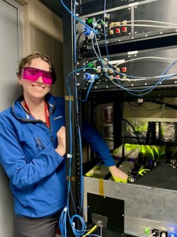 A woman standing next to an equipment rack that holds the atomic clock technology