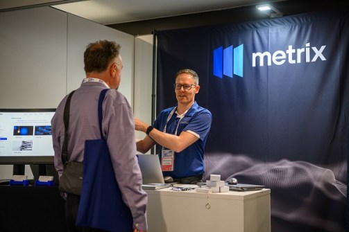Matt Robinson from Chartis Technology standing behind a counter at a trade exhibition, speaking with a visitor about Metrix software