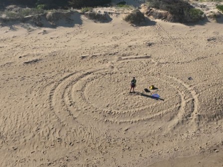 A drone's eye view of a woman standing on a beach, controlling the drone, while undertaking mapping of Victorian beaches
