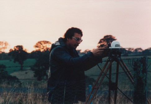 A man adjusting a GPS antenna on top of a tripod, in a country field