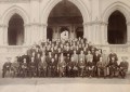 A sepia photo of a large group of people in old-fashioned dress, posing as a group on the steps of a standstone building