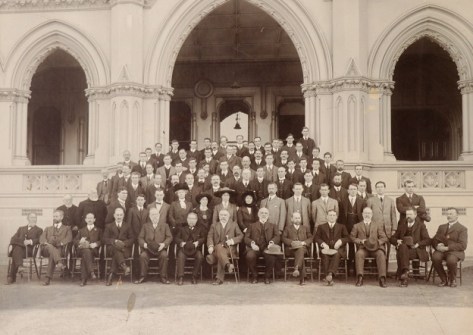 A sepia photo of a large group of people in old-fashioned dress, posing as a group on the steps of a standstone building
