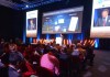 Looking from behind at an audience at a conference, with a person speaking on-stage in the background