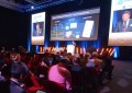 Looking from behind at an audience at a conference, with a person speaking on-stage in the background