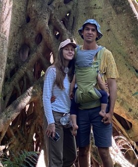 A man, woman and baby standing next to a large tree trunk