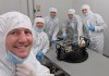A group of people in lab clothing inside a cleanroom, with a SWIRSAT satellite on a table