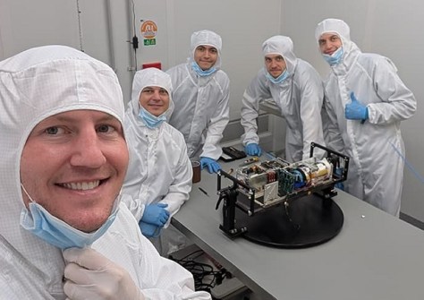 A group of people in lab clothing inside a cleanroom, with a SWIRSAT satellite on a table