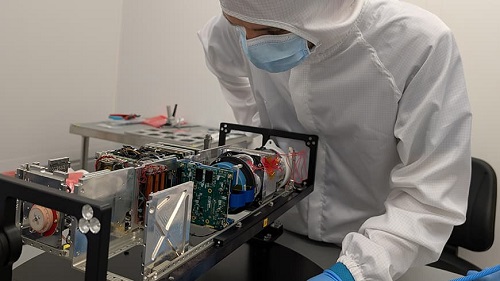 A man in lab clothing inside a cleanroom, with a SWIRSAT satellite on a table