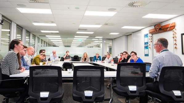 About a dozen people sitting around a square-configured group of desks, discussing Landonline