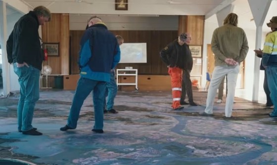 Six people standing on a Big Map, inspecting it, inside a community hall
