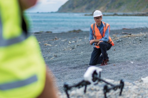A man crouching on a rocky beach, with a drone in front of him