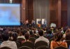 A blurred view of a conference hall, taken from the back, overlooking audience heads