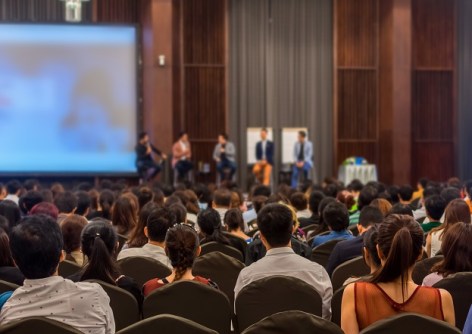 A blurred view of a conference hall, taken from the back, overlooking audience heads